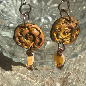 May include: A pair of dangle earrings with a yellow and brown floral design. The earrings are made of metal and have a small, clear crystal bead at the bottom.