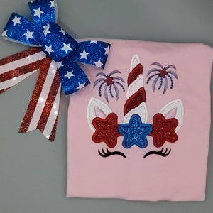 May include: A pink t-shirt with an embroidered unicorn design. The unicorn has a red, white, and blue horn and stars. The design also includes fireworks.