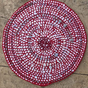 May include: A round, red and white braided rug made from recycled materials. The rug has a concentric circle pattern.