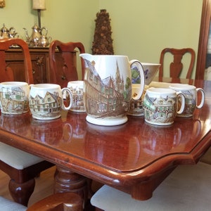 May include: A set of six vintage ceramic mugs and a pitcher with a scene of a quaint English village. The mugs and pitcher feature a brown, green, and white color scheme with a detailed illustration of buildings and trees.