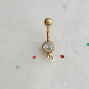 May include: A gold-coloured belly button ring. It features a clear, round gemstone at the bottom and a curved barbell design. The ring has a small ball at the top and a loop at the bottom for attaching charms.