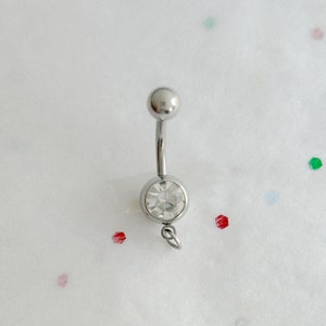 May include: A silver-colored belly button ring with a clear, round gemstone at the bottom and a small ball at the top. The ring is curved and has a small loop attached to the gemstone setting. The background is white with scattered colorful confetti.
