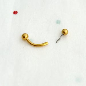 May include: A gold-coloured curved barbell with a ball on each end. The barbell is separated from the ball, showing the post.