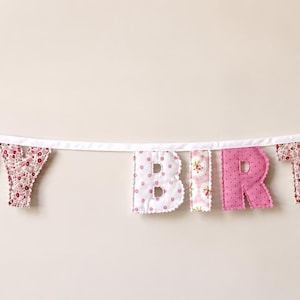 May include: A fabric banner spelling out the words HAPPY BIRTHDAY in individual letters. The letters alternate between a pink floral pattern and a solid pink fabric. The letters are attached to a white ribbon with white bows at each end.