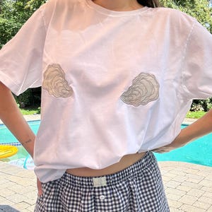 May include: A white short-sleeved t-shirt featuring two embroidered oyster shell designs on the chest area. The shirt is paired with navy blue and white gingham patterned shorts that have an elastic waistband and a single center button.