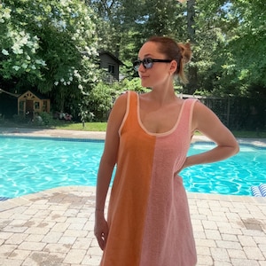 May include: A sleeveless terry cloth dress featuring a vertical color-blocked design with one half in bright orange and the other half in soft pink. The garment has a scoop neckline and light-colored trim along the edges of the armholes and neck.