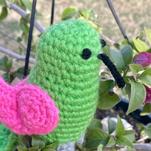 May include: A crocheted hummingbird toy with green and pink yarn. The toy has a long black beak and black eyes.