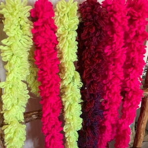 May include: A display of colorful, textured yarn garlands in shades of neon green, hot pink, deep red, and purple. The garlands are hanging from a wooden structure, showcasing their fluffy, looped texture. The vibrant colors create a visually striking display.