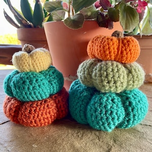 May include: Two handmade crochet pumpkins in shades of orange, teal, green, and yellow. The pumpkins are stacked, with a small brown stem on top. The pumpkins are displayed on a stone surface, with potted plants in the background.