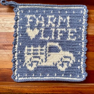 May include: A square, blue and white crocheted potholder with the words "FARM LIFE" and a truck design. The potholder has a scalloped edge and a loop for hanging. The design is in white against a blue background.