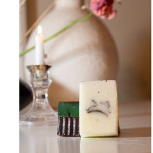 May include: Three bars of handmade soap, one is white with a green leaf design, one is green, and one is brown with white stripes.