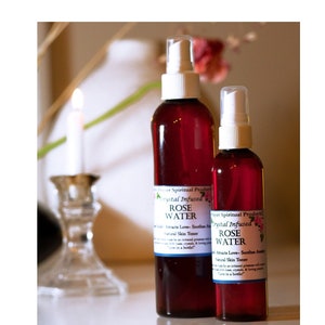 May include: Two bottles of rose water, one large and one small, with white spray tops. The bottles are filled with a deep red liquid and have labels that read "Crystal Infused Rose Water". The bottles are on a reflective surface.