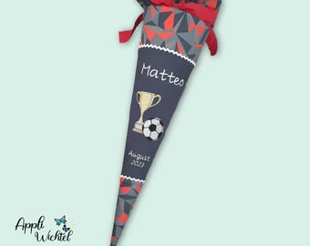 May include: A cone-shaped school gift bag featuring a dark grey fabric body with a gold trophy and football graphic. The name Matteo and the text August 2023 are embroidered on the front. The top has a red and grey geometric fabric closure.