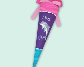 Puede incluir: Una bolsa de regalo escolar en forma de cono con cuerpo morado, una ilustración de un delfín y el nombre Mia impreso en blanco. La parte superior tiene un cuello de tela rosa y un lazo a juego. La parte inferior muestra olas azules.