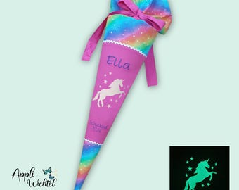Puede incluir: Un cono escolar cónico con un cuerpo de tela rosa y una sección superior de colores arcoíris atada con una cinta rosa. El cono muestra el nombre bordado Ella, una silueta de unicornio blanco y el texto Schulkind 2024. Un recuadro pequeño muestra al unicornio brillando.