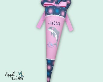 Puede incluir: Un cono escolar cónico con una sección central rosa que lleva el nombre Julia y un gráfico de un delfín gris. Las secciones superior e inferior son azul oscuro con patrones circulares rosas y azules. Una cinta rosa cierra la parte superior.