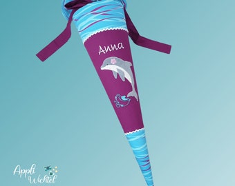 Puede incluir: Una bolsa de regalo de tela en forma de cono con cuerpo morado, un gráfico de delfín y el nombre Anna en texto blanco. Las secciones superior e inferior son azul claro con líneas onduladas azul oscuro, con un lazo morado oscuro en la parte superior.