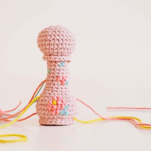 Puede incluir: Un sonajero hecho a mano de ganchillo en un tono rosa suave. El sonajero presenta una parte superior redondeada y una base bulbosa, adornada con detalles florales bordados en colores pastel. Hilos de colores se extienden desde la base, agregando un toque lúdico a este juguete artesanal para bebés.