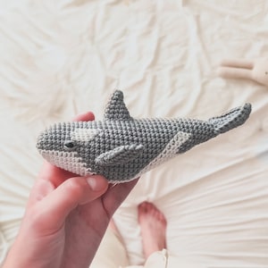 Puede incluir: Un juguete de orca de crochet gris y blanco. El juguete está siendo sostenido por una mano.
