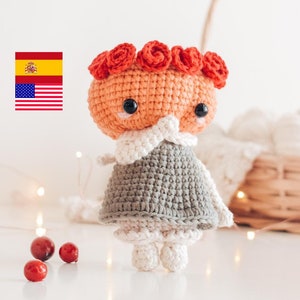 Puede incluir: Una muñeca de crochet con la cabeza de color melocotón y un vestido gris. La muñeca tiene una corona de flores rojas y ojos negros. La muñeca está de pie sobre una superficie blanca.