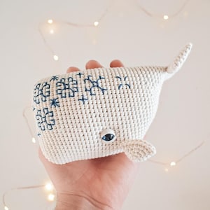 May include: A white crocheted whale with blue embroidered details. The whale has a single blue eye and a small, crocheted tail.