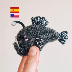 Puede incluir: Un juguete de peluche de pez pescador negro tejido a crochet con detalles plateados. El juguete está siendo sostenido por una mano.