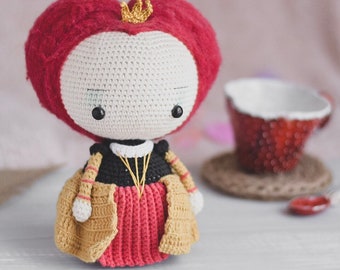 May include: A crocheted doll with a red heart-shaped head, a gold crown, and a red and gold dress. The doll is sitting on a white surface.