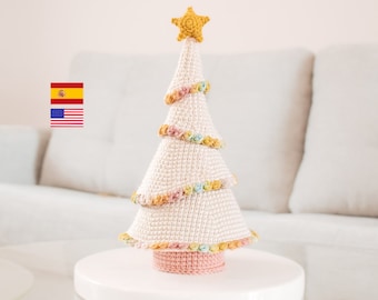 Op de afbeelding: Een witte gehaakte kerstboom met een roze basis en een gele ster bovenop. De boom is versierd met kleurrijke gehaakte bloemen.