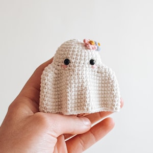 May include: A white crocheted ghost with black eyes and pink cheeks. The ghost has a small flower on its head with pink, yellow, and blue petals.