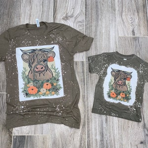 Pode incluir: Duas camisetas verdes com um gráfico branco de uma vaca das Terras Altas com folhagem de outono e abóboras. A camiseta maior tem um visual desgastado com respingos de tinta branca.