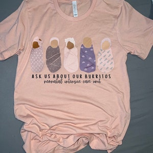 Op de afbeelding: Een roze T-shirt met een afbeelding van vijf gewikkelde baby's en de tekst "Vraag ons naar onze burrito's neonatale intensive care unit".