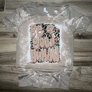 May include: A beige t-shirt with a bleached design featuring the text "Be a good human" in a brown and orange colour scheme with a leopard print background.