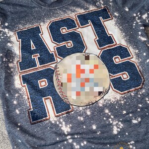 Könnte beinhalten: Ein graues T-Shirt mit einem Bleiche-Spritzer-Design, das das Wort "ASTROS" in großen, blauen Buchstaben mit einem Baseball in der Mitte zeigt. Der Text "ALEJANDRA Creations" ist in der rechten unteren Ecke sichtbar.