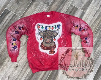 May include: Red sweatshirt with a white and red Highland cow wearing a red and black scarf with a string of colorful Christmas lights above its head. The sweatshirt has a white splatter pattern.