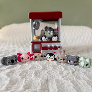 May include: A collection of small crocheted animal plushies, including a koala, axolotl, owl, penguin, bunny, cat, and a green creature. The plushies are arranged in a line on a white fluffy surface. A red and white claw machine is in the background.