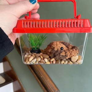 May include: A small, clear plastic terrarium with a red lid and handle. Inside, there is a brown and black crocheted animal, green plastic plants, and small brown rocks.