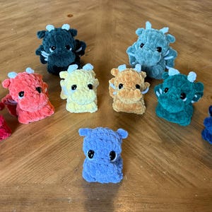 May include: A collection of nine crocheted plush dragon toys in various colours, including red, orange, yellow, green, blue, and purple. The dragons have white eyes and are sitting on a wooden surface.
