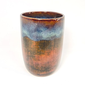 May include: A handcrafted ceramic tumbler with a unique, textured surface. The cup features a gradient of colours, transitioning from a deep brown at the base to a blue-green hue at the rim. The design is reminiscent of natural stone.
