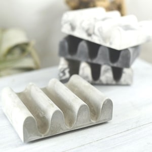 May include: A stack of four soap dishes in shades of gray and white. The front-most dish is light gray with three curved ridges. The other dishes are stacked behind it, with a marble-like pattern. The dishes are made of a solid material and sit on a white surface.