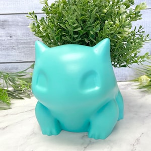 Peut inclure: Un pot en céramique turquoise en forme de personnage de dessin animé avec de grands yeux et un corps rond. Le pot est rempli de plantes vertes feuillues.