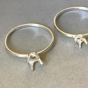 May include: Two silver ring settings with a pronged design, ready for stone placement. The rings are circular, with a polished finish, and sit on a grey surface. The settings are designed to hold a gemstone.
