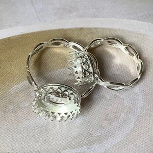 May include: Close-up of three silver-colored ring settings. The settings have a decorative crown design around the edges. The rings have an open design, with a decorative pattern on the band.