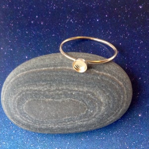 May include: A silver-colored ring with a round bezel setting. The ring is resting on a smooth, gray stone against a blue background with white speckles. The ring's band is thin and circular.