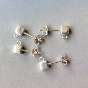 May include: Five silver stud earrings with a square design and a small loop at the bottom. The earrings have a post and butterfly backing. The earrings are displayed on a grey surface.