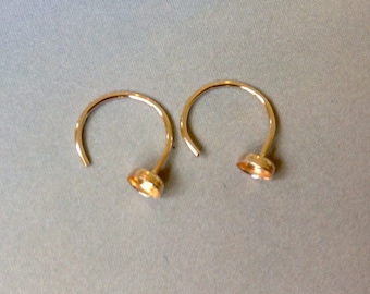 May include: Two gold-colored hoop earrings with a post and a small, round setting. The earrings are displayed on a light gray surface. The earrings have a simple, elegant design.