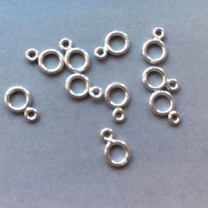 May include: A selection of small, silver-coloured jewellery clasps. Each clasp has a circular ring attached to a smaller ring. The clasps are spread out on a grey surface, with soft, diffused lighting.