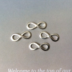 May include: Four silver infinity charms are shown on a grey background. Each charm is in the shape of the infinity symbol, with a smooth, polished finish. These charms are likely used for jewellery making or crafting projects.