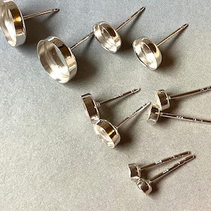 May include: A collection of silver-colored stud earring components, including posts and bezel settings. The components are arranged on a light gray surface, showcasing their reflective, polished finish. The earrings are designed for jewelry making.