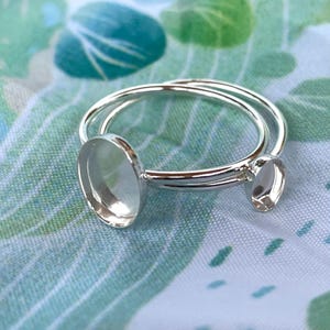 May include: A silver ring with two oval settings. The ring features a double band design and is set against a green and blue patterned background. The settings are empty, ready for gemstones or other embellishments.