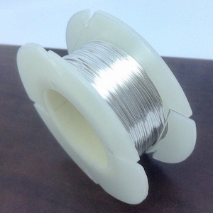 May include: A white plastic reel holds a tightly wound coil of silver-coloured wire. The reel has a simple design with slots for the wire to pass through. The wire appears to be made of a shiny metal.